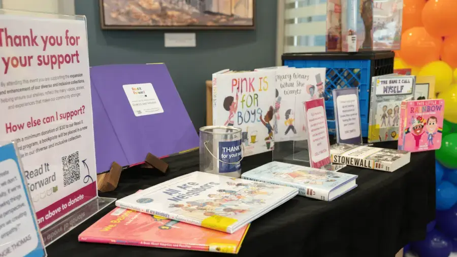Display table featuring diverse and inclusive library books, donation information, and a Read It Forward fundraising campaign, with colorful balloons in the background.