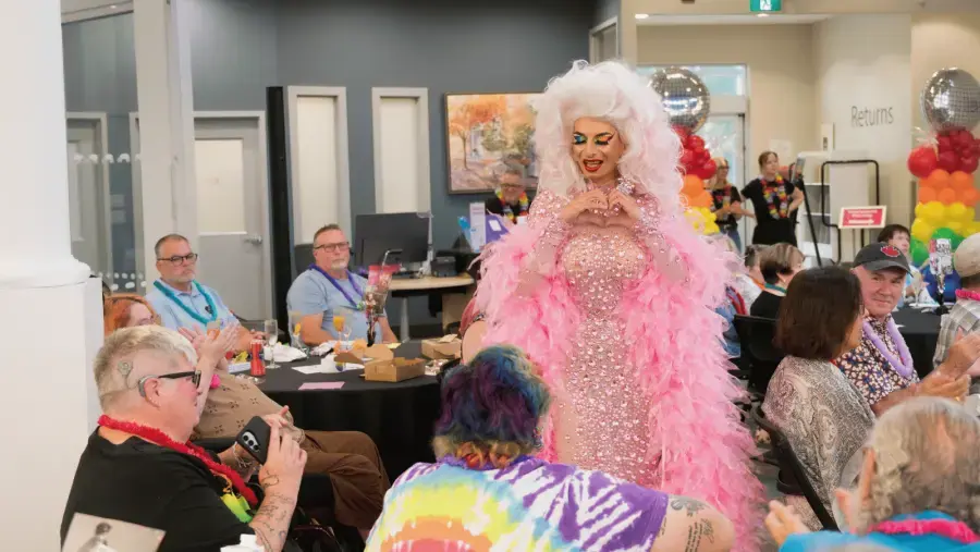 A performer wearing a sparkling pink outfit and feathered wrap walks among guests seated at tables during event.