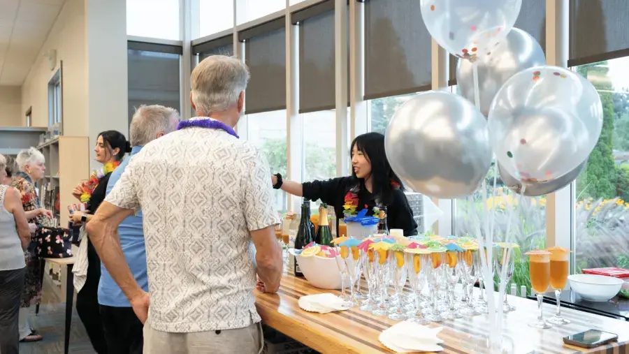 Attendees gather around a refreshment table decorated with silver and confetti-filled balloons, while sparkling beverages are being served.