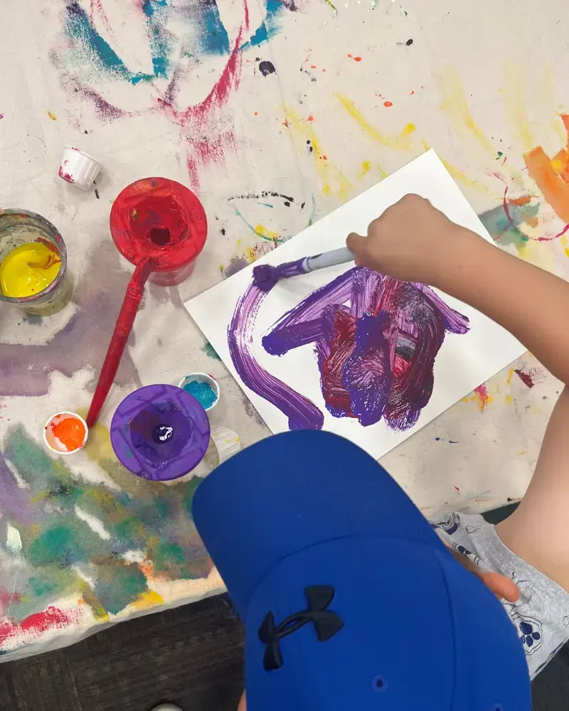 A child wear a blue baseball cap is painting on white paper with purple paint. There are three paint pots on the table with red, purple and yellow paint. The photo is a birds eye view of the table, and the table is covered with a drop cloth covered in paint splashes.