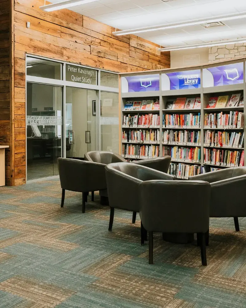 Photo of the interior of the NOTL Library. There are two seating groups with three grey vinyl chairs at each. They are in front of a large shelf of books with branding at the top of the shelves. The background shows the library's quiet room, enclosed behind glass doors and surrounded with a wall of reclaimed wood.