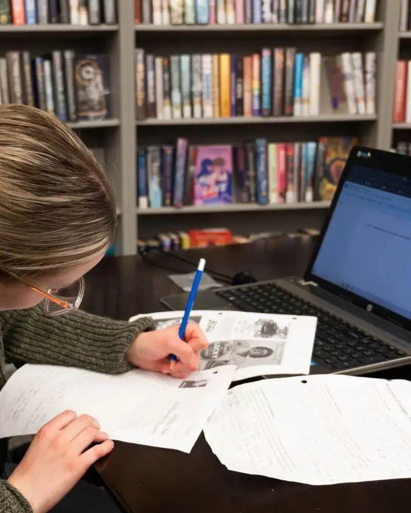 Photo of a young woman working on a project at a table in the library. She has a laptop and a number of papers in front of her. In the background are a series of full bookshelves.
