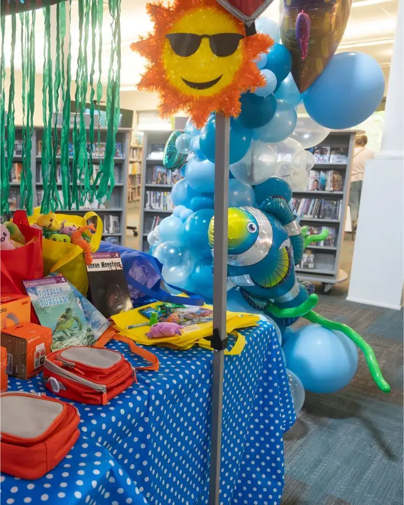 Photo of a table with a blue polka-dot tablecloth. There are colourful books and toys on the table. At the top centre is a yellow and orange paper sun wearing sunglasses. In the background is a balloon sculpture in blue-toned balloons with colourful fish balloons throughout.