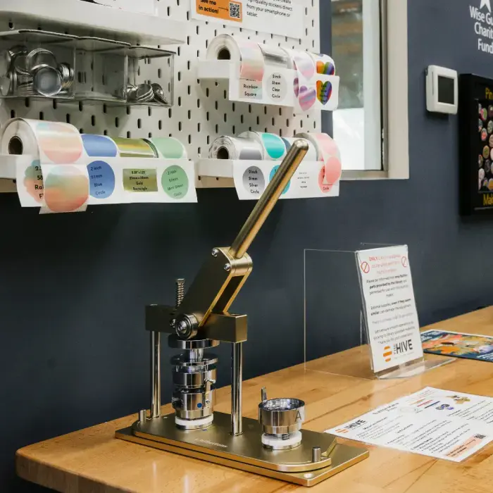 Image of a button maker device, made of a gold-toned metal. It is sitting on a light wood work table. In the background is a pegboard with sticker rolls and craft supplies attached.