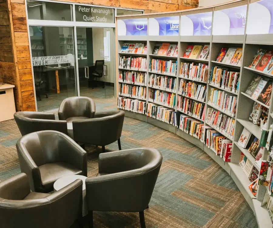 Photo of NOTL Library interior featuring grey vinyl chairs arranged in sets of three in front of a large colourful bookshelf. There is a window and sliding glass doors in the background leading to the Quiet Room.