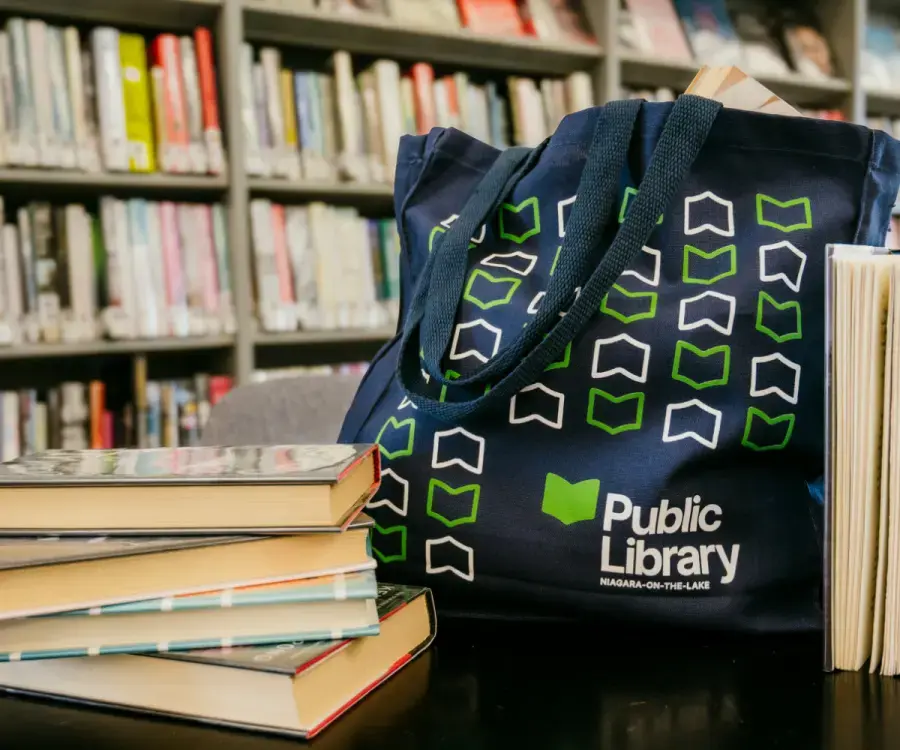 Image features a navy blue NOTL Library tote bag featuring the library's book icon logo with a stack of four books directly beside it to the left. Behind the bag is a large bookshelf filled with library books.