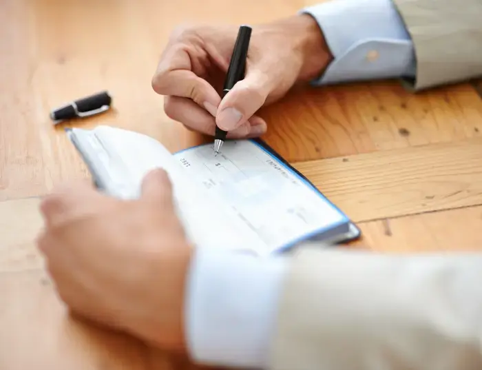Image shows a man's hands; he is wearing a beige suit jacket with light blue shirt cuffs below. He holds a pen and is writing in a chequebook.