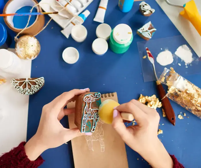 Birds-eye view photo of a craft table with a blue tablecloth and a number of crafting supplies including paint, glitter and a paint knife. There are two hands applying paint to a stamp with a foam applicator. The stamp is in the shape of a reindeer.