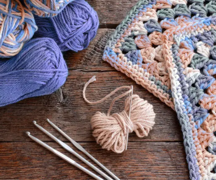 Photo shows a crochet scene. A few balls of purple and pink yarn with three crochet hooks are on the right side. On the left are a couple of crochet squares that have been made with the yarn.
