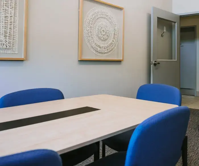 The Commons, a room that is available to rent at the library. There are two large framed art pieces on the wall that are geometric patterns created with paper. In the foreground is a light wood coloured table with chairs in blue upholstery. A light grey door with a large window opens into a hallway in the background.