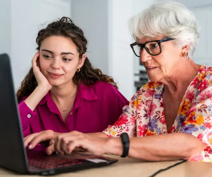 Photo of two women sitting at a desk, one young, one a senior citizen. They are both looking at a computer screen. The older woman has her hands on a keyboard.
