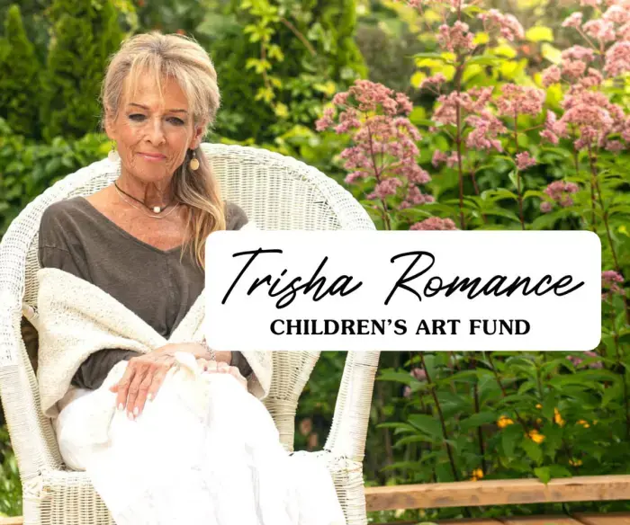 A photo of the artist Trisha Romance. She is outside, surrounded by green bushes and pink flowers, sitting on a large, white, woven chair. The text in the foreground reads 